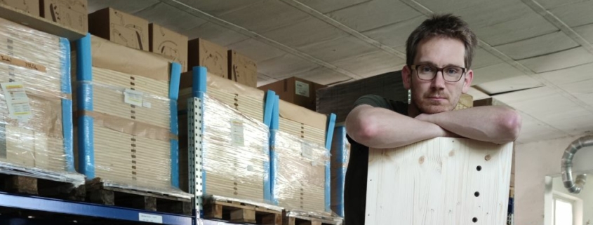 David Witzeneder hält Holz-Seitenteil einer Wurmkiste in der Hand während er auf Holzstoß in der Wurmkistenwerkstatt sitzt.